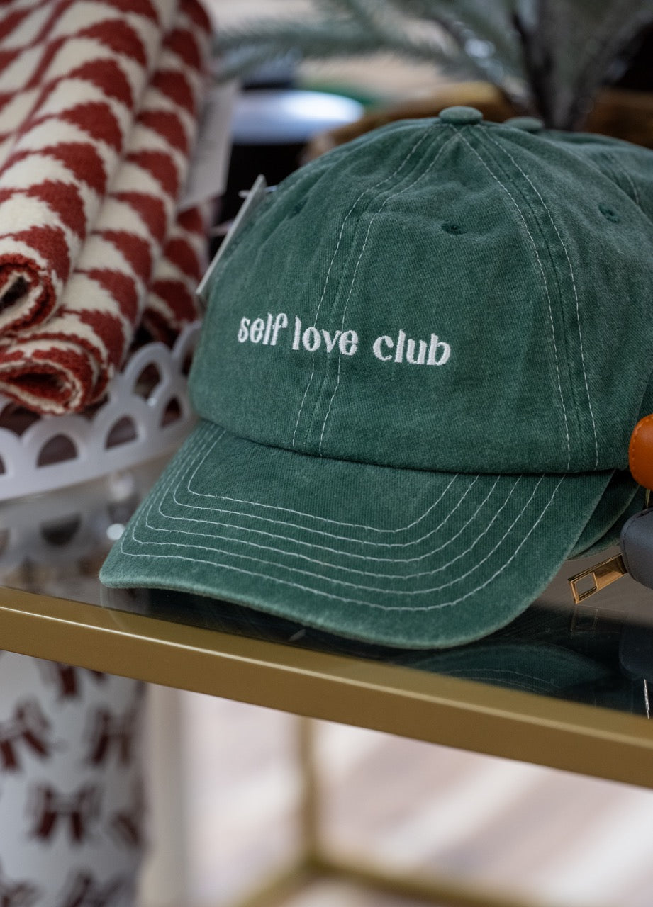 Green cap with 'self love club' text on a blurred background